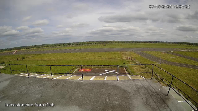 A view from a webcam showing an open grassy area with a runway or taxiway extending into the distance. The sky is partly cloudy with gray and white clouds. There is a fenced area in the foreground, and the label "Leicestershire Aero Club" is displayed in the bottom left corner. The timestamp in the top right corner reads "07-05-2025 13:00:22".