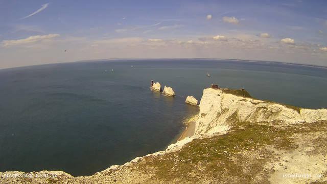 A scenic view of white chalk cliffs along the coastline with a lighthouse on top of one of the cliffs. The cliffs extend into the ocean, with several large rock formations rising from the water nearby. The sky is partly cloudy with a few contrails, and several sailboats are visible on the calm sea.