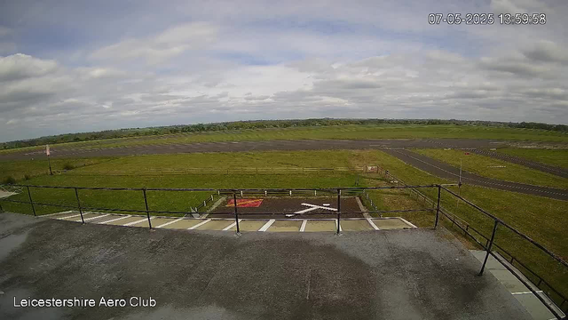 A view from a webcam at Leicesterhire Aero Club shows an open grassy airfield with a runway stretching into the distance under a cloudy sky. In the foreground, there are parking spaces and a small concrete area with a metal railing. The date and time displayed are July 5, 2025, at 13:59:58.
