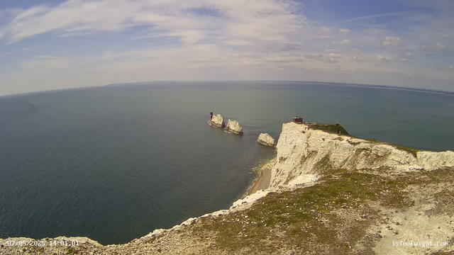 A panoramic view of tall white chalk cliffs extending into the sea, with some sea stacks near the shoreline. The sky above is partly cloudy with wispy clouds, and the water below is calm, reflecting the sky. Atop the cliffs, there is a small building and a lighthouse on one of the stacks. The image has a timestamp in the bottom left corner.