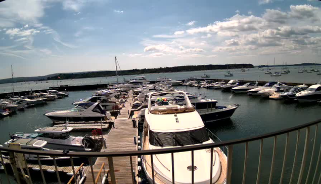A marina with numerous boats and yachts docked along wooden piers. The water is calm, and there are sailboats in the distance on a partly cloudy sky. A metal railing is visible in the foreground.