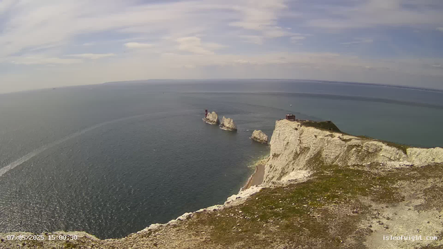 A view of white chalk cliffs surrounded by the ocean, with several large rock formations rising from the water. There is a small building and a lighthouse on the edge of the cliff, and a sailboat is visible in the distance. The sky is partly cloudy. The scene appears to be taken from a webcam overlooking the coastline.