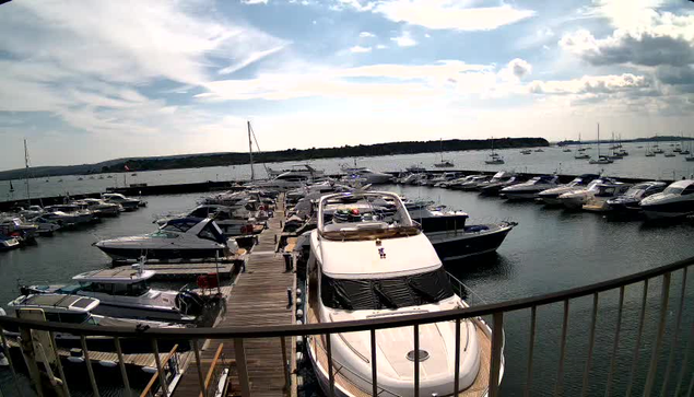 A marina filled with various boats and yachts docked along wooden piers, with a calm body of water extending into the distance and a partly cloudy sky overhead. A metal railing is visible in the foreground.