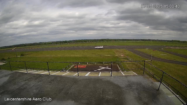 A small aircraft is taxiing on the runway under a cloudy sky. The photo is taken from an elevated viewpoint, possibly from a control tower or observation deck. The runway is surrounded by green grass, and there are some parked vehicles and equipment in the foreground. The text "Leicestershire Aero Club" is visible in the bottom left corner, and the timestamp in the top right corner reads "07-05-2025 15:00:40."
