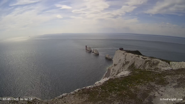 A view of the white cliffs and the sea during daytime, with three prominent rock stacks near the shoreline, one of which has a lighthouse on top. The sky is partly cloudy, and the water reflects the sunlight. There is a small structure on the highest point of the cliff. The image has a timestamp in the lower left corner and a website address in the lower right corner.