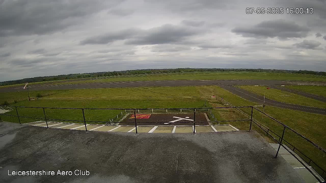 The image shows an outdoor view from a webcam at Leicestershie Aero Club. In the foreground, there is a rooftop area with a black railing and a small section of marked parking spaces. Below, there's a patch of grass with a large white "X" painted on the ground. Beyond the grassy area, there's a paved runway stretching into the distance under a cloudy sky with dark gray clouds. The landscape is flat with distant trees and fields visible on the horizon. The timestamp in the top right corner reads "07-05-2025 16:00:13."