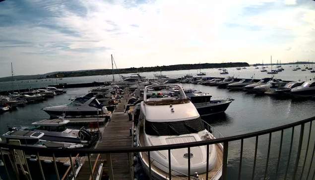 A view from a balcony overlooking a marina filled with various boats and yachts docked along wooden piers, with a cloudy sky overhead and distant land on the horizon.