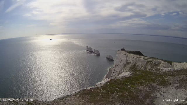 A panoramic view of white chalk cliffs overlooking the ocean, with several large sea stacks and a lighthouse on the edge of the cliff. The sky is partly cloudy, and the sunlight reflects off the water's surface. There is a small building or structure near the lighthouse. The photo appears to be taken from a webcam at the top of the cliffs.