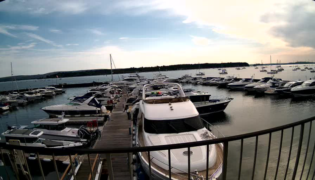 A marina filled with numerous boats and yachts docked along wooden piers, with calm waters reflecting the vessels and a partly cloudy sky overhead.