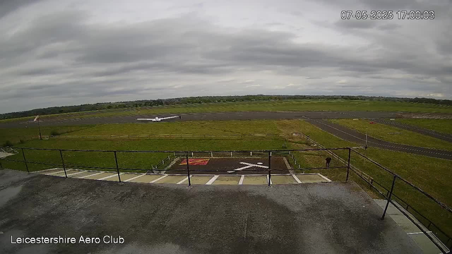 A wide view of an airfield on a cloudy day, with the runway extending into the distance. In the foreground, there is a balcony with a metal railing and a marked helipad area. A small aircraft is parked near the runway, and a person is walking along the grass. The image has a timestamp in the top right corner showing July 5, 2025, at 17:00:03, and "Leicestershire Aero Club" text is visible at the bottom left corner.