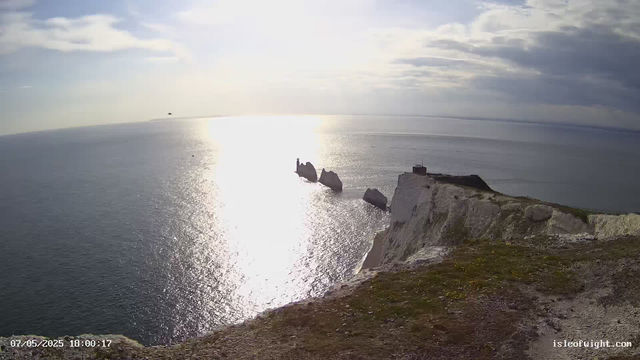 A view of a rugged white chalk cliff overlooking a calm ocean. The sun is low, creating a bright reflection on the water. In the distance, several large rock formations and a small lighthouse or tower perched on one of the rocks are visible. The sky is partly cloudy with patches of sunlight breaking through. The photo appears to be taken from a high vantage point on the edge of the cliff.