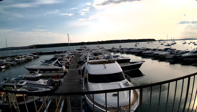 A marina filled with numerous boats and yachts docked along wooden piers, with calm water reflecting the vessels and the sky overhead. The sky is partly cloudy with a warm, golden glow suggesting either sunrise or sunset. A curved railing is visible in the foreground.