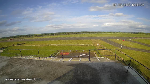 An outdoor view from a webcam at Leicester Shire Aero Club shows a partly cloudy sky over a grassy airfield. In the foreground, there is a parking area with a few marked parking spaces and a helipad with a white "H" and a red border. Surrounding the airfield is a fence and a small flagpole with a flag. The horizon features a line of trees and open countryside in the distance. The timestamp in the top right corner indicates the date and time as July 5, 2025, at 6:00 PM.