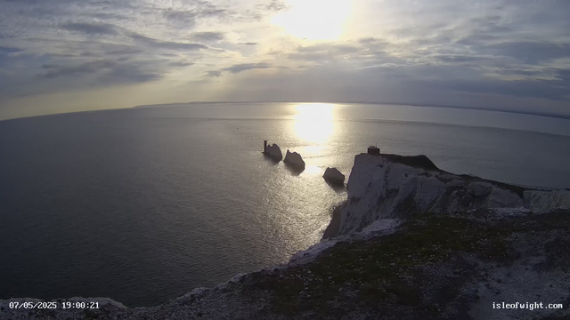 A panoramic view of a coastal landscape during sunset or sunrise, with the sun partially obscured by clouds. The image features white chalk cliffs with a small structure at the top right, and several sea stacks rising from the water near the center. The water is calm, reflecting the light from the sky. The scene has a tranquil and scenic atmosphere.