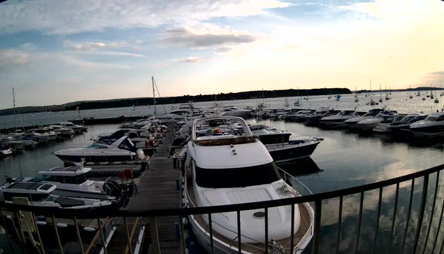 A marina filled with numerous boats and yachts docked along wooden piers, with a calm water surface reflecting the vessels. The sky is partly cloudy with the sun either rising or setting, casting a warm glow over the scene. A black railing is visible in the foreground.
