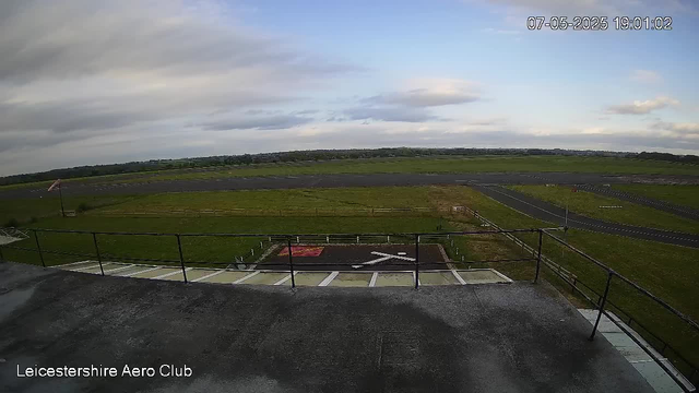 The image shows an empty runway and grassy airfield area viewed from an elevated position. There is a fenced section on the ground with a painted white arrow and a red emblem. In the background, the sky is partly cloudy with patches of blue, and the horizon features flat land stretching into the distance. The timestamp on the image reads "07-05-2025 19:01:02," and there is a label at the bottom left that says "Leicestershire Aero Club."