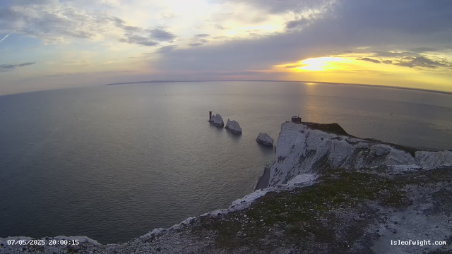 A view from a webcam showing white chalk cliffs overlooking the sea at sunset. Several large rock formations are visible in the water just off the coast, along with a lighthouse and a small building on top of the cliffs. The sky is partly cloudy with the sun low on the horizon, casting a golden light across the scene. There is a timestamp in the bottom left corner, and the website isleofwight.com is visible in the bottom right corner.