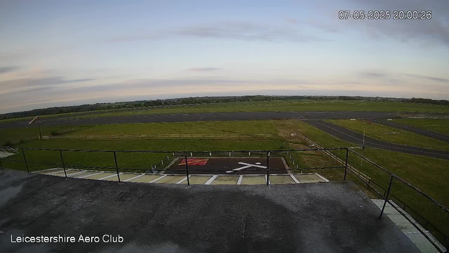An outdoor view from a webcam showing a runway and grassy fields extending into the distance under a partly cloudy sky during early evening. The timestamp indicates it is 20:00 on July 5, 2025, and the location is Leicester Shire Aero Club. The foreground includes a safety helipad with markings and a fence, with a ramp leading down to the ground. No people or aircraft are visible.