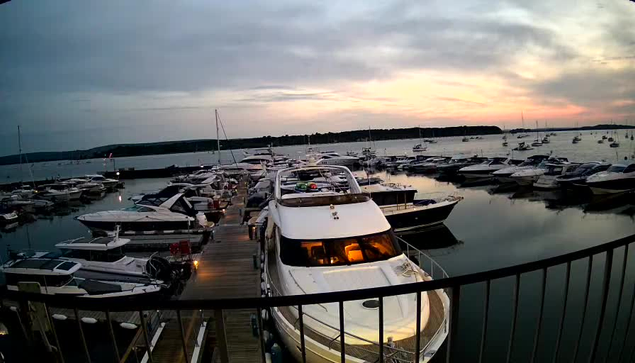 A marina filled with numerous boats and yachts docked at a wooden pier during sunset. The sky is partly cloudy with a mix of dark and light clouds, and the water reflects the boats and the fading light of the sunset. In the foreground, there is a black railing along the edge of the dock.