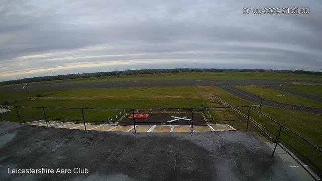A wide view from a webcam showing an empty airport tarmac and runway beneath a cloudy sky. In the foreground, there is a helipad marked with a large white "H" and a red square. Surrounding the area are grassy fields extending into the distance. The image has a timestamp in the upper right corner indicating it was taken on July 5, 2025, at 21:01. At the bottom left, the text reads "Leicestershire Aero Club."