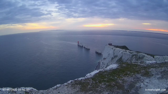 A view from a webcam showing white chalk cliffs overlooking the ocean during sunset. The sky is partly cloudy with some orange and yellow hues near the horizon. In the water near the cliffs, there are two boats, one with a red and white lighthouse. The date and time stamp in the bottom left corner reads "07/05/2025 21:01:09," and a website URL "isleofwight.com" is visible in the bottom right corner.