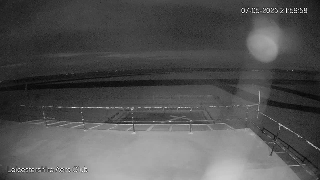 A black-and-white image from a webcam showing an empty aircraft parking area at night. The top right corner displays the date and time as 07-05-2025 21:59:58. There are a few light reflections and a faint glow in the sky, with a barrier or railing visible around a central circle. The bottom left corner reads "Leicestershire Aero Club."