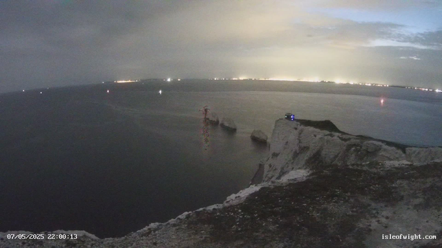 Nighttime view of a coastal landscape with white chalk cliffs in the foreground. The sea is calm, with a few rocks visible in the water near the cliffs. In the distance, faint city lights and illuminated structures line the horizon, and the sky is partially cloudy with a soft glow from the city lights. A small lighthouse or beacon is visible on top of the cliffs. The timestamp indicates the photo was taken on May 7, 2025, at 22:00.