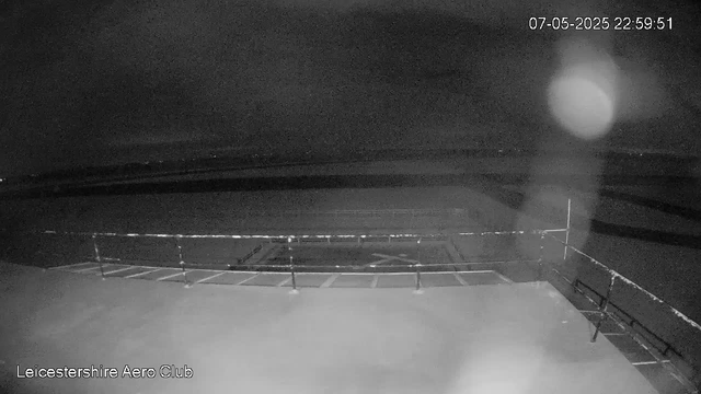 A black and white nighttime scene of a parking lot with empty parking spaces enclosed by a metal railing. The sky appears cloudy or foggy, with a large bright moon or light source partially obscured in the upper right. The timestamp in the upper right corner reads "07-05-2025 22:59:51," and the bottom left corner displays "Leicestershire Aero Club."