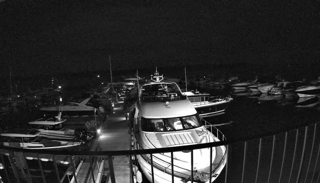 A night scene at a marina showing multiple boats docked along wooden piers. The boats vary in size and include motor yachts and smaller vessels, all moored closely together. In the foreground, there is a railing, and the water is calm, reflecting the boats and faint light from distant sources. The sky is dark with no visible stars or moon.