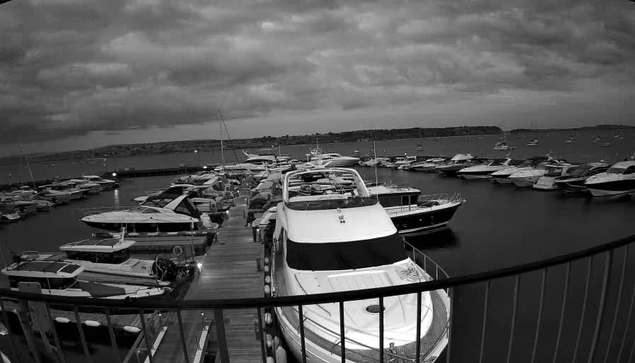 A black-and-white image of a marina filled with numerous boats and yachts docked along wooden piers. The photograph is taken from a balcony or platform with a curved railing in the foreground. The sky above is partly cloudy, giving the scene a moody, overcast atmosphere. In the background, there is water extending towards the horizon with more boats visible in the distance.