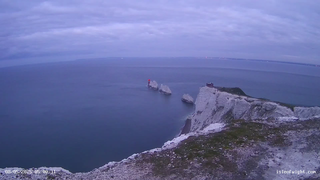 A view of white chalk cliffs extending into the ocean, with several large rock formations near the water's edge. One of the rocks has a small lighthouse with a red light on top. The sky is mostly cloudy, and the timestamp shows the date as August 5, 2025, at 05:00:31. The URL "isleofwight.com" is visible in the bottom right corner.