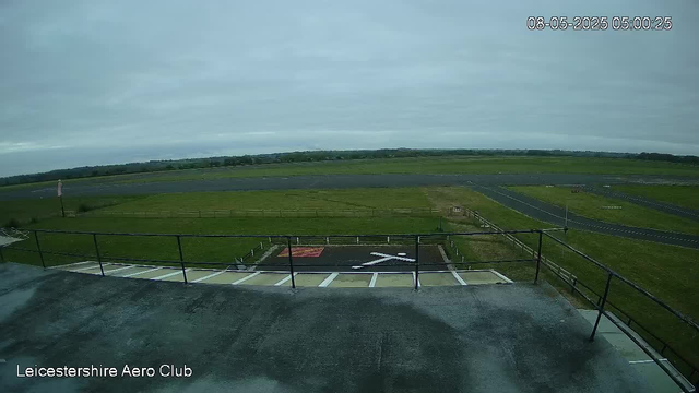 An empty helipad with a white helicopter symbol painted on it, surrounded by a black safety railing. To the left, there is a small statue or figure. The scene overlooks a wide grassy field and an airstrip in the distance. The sky is overcast with grey clouds, and the timestamp shows the date and time as August 5, 2025, at 05:00:25. The bottom left corner has the text "Leicestershire Aero Club."