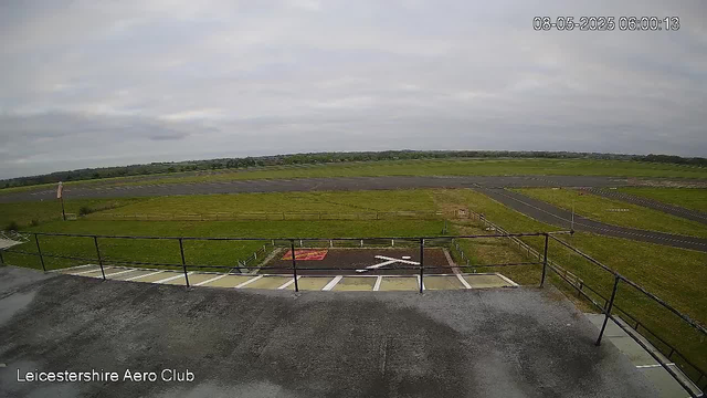 A view from a webcam at Leicestershire Aero Club shows an overcast sky and a runway extending into the distance. In the foreground, there is a small helipad with a white helicopter symbol and a red marking, surrounded by a metal railing. The landscape is mostly flat with green grass and some paved areas, and there are a few distant structures or trees visible on the horizon. The timestamp on the image indicates it was taken on August 5, 2025, at 6:00 AM.