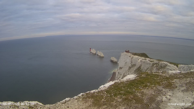 A wide view of white chalk cliffs rising from the ocean, with a small building and a lighthouse on one of the cliffs. The sea is calm and stretches to the horizon under a cloudy sky. The image is shot from a high vantage point on the cliff edge.