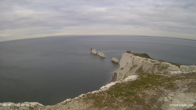 A view of white chalk cliffs along a coastline with a grassy top, extending into the calm ocean under a cloudy sky. A small building is situated at the edge of the cliffs, and large rocks are visible in the water near the base of the cliffs. The image includes a lighthouse or navigational tower on a rock formation offshore, with an overcast sky overhead.