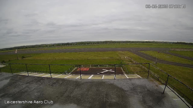A view from a webcam at Leicesterhire Aero Club shows an empty tarmac area with a helicopter landing pad marked with a white 'H'. Surrounding the tarmac are green grassy fields, with a paved road curving across the landscape. The sky is overcast with gray clouds, and the scene is calm and empty. The timestamp in the top right corner indicates it was taken on August 5, 2025, at 7:00 AM.