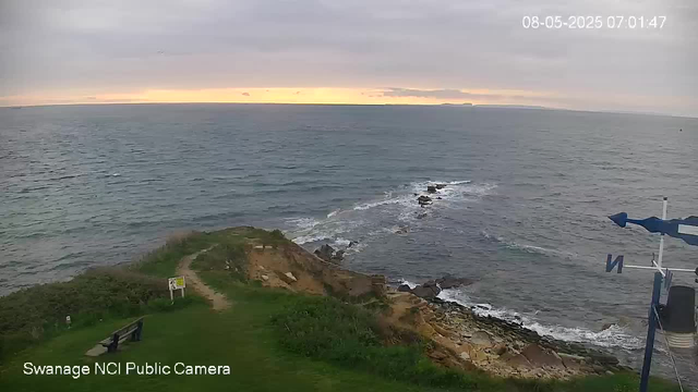 A view of the coastline from a webcam, showing a grassy area with a bench in the foreground, a rocky shoreline extending into the ocean, and an overcast sky at sunrise or sunset in the background. The timestamp shows the date as August 5, 2025, early morning. The camera label indicates it is the Swannage NCI Public Camera.
