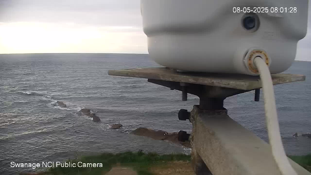 A webcam captures an ocean view with a rocky shoreline. In the foreground, there is a white device mounted on a metal bracket, connected by a white cable. The sky appears overcast, with a few clouds, and the water looks relatively calm. The image has a timestamp in the upper right corner showing "08-05-2025 08:01:28", and text in the lower left corner reads "Swanage NCI Public Camera."