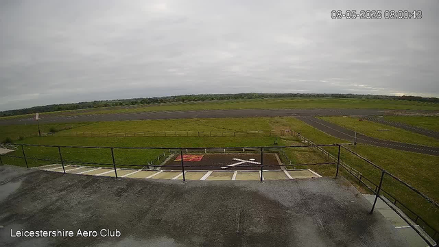 An overcast sky covers the open landscape of Leicestershire Aero Club, with a long runway stretching across the horizon. In the foreground, there is a small parking area with painted lines and an area marked with a large white airplane symbol. A metal railing runs along the edge of a concrete platform, which appears to be an observation deck or viewing area. The scene is quiet and empty, with no visible aircraft or people. The timestamp in the upper right corner indicates the image was captured on August 5, 2025, at 8:00 AM.