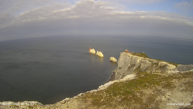 A view from a webcam showing white chalk cliffs extending into the ocean, with large rock formations just offshore. There is a small building on top of the cliffs, and a red and white lighthouse on one of the rocks in the water. The sky is partly cloudy with patches of blue. The timestamp at the bottom left indicates the date and time as 08/05/2023 at 08:01:03.