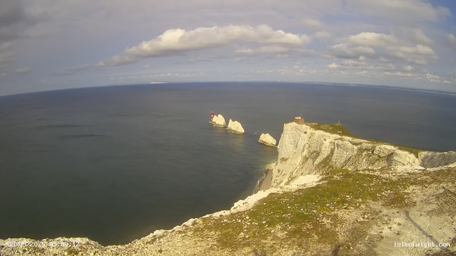 A panoramic view of white chalk cliffs rising out of the ocean, with a prominent cliff featuring a lighthouse at the top. Several large rock formations are visible offshore, including one with a red and white lighthouse. The sky is partly cloudy with patches of blue, and the water appears calm with gentle waves. The scene captures a coastal landscape during daytime.