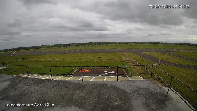 An overcast sky with grey clouds over a wide open grassy area at Leicestershre Aero Club. There is a small helipad marked with a white helicopter symbol and a large white "X" in the center. A section of paved parking with white lines and a black railing in the foreground. The date and time display reads "08-05-2025 09:00:33" in the upper right corner.