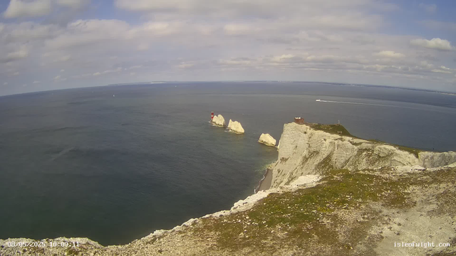 A panoramic view of white chalk cliffs along the coastline with a lighthouse on one of the cliffs, four large chalk stacks in the sea nearby, and a small building on the edge of the cliff. The ocean is calm, and a boat is visible moving across the water, leaving a white trail behind. The sky is partly cloudy with patches of blue.