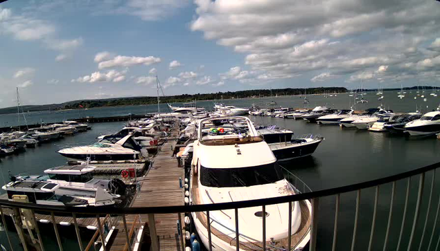 A marina filled with various boats docked along wooden piers, with a railing in the foreground. The sky is partly cloudy, with some fluffy clouds scattered across a blue sky. The water is calm, reflecting the boats and the sky. In the background, there is a landmass with trees.