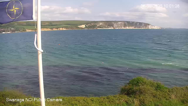 A view of a coastal landscape with a blue flag featuring a yellow compass rose emblem in the foreground on the left. The image shows a calm sea with gentle waves, a green grassy area with some bushes at the bottom, and white cliffs in the distance under a partly cloudy sky. The timestamp in the top right corner reads "08-05-2015 10:01:28."