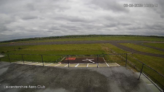 An empty airport parking area with several designated parking spots, a small red object, and a black arrow painted on the tarmac indicating direction. Beyond the parking area, there is a grassy field with a few small tracks or paths, and an open sky with thick, gray clouds. In the background, there are low hills or distant trees. The image is timestamped "08-05-2025 10:00:48" in the upper right corner, and "Leicestershire Aero Club" is written in the lower left corner.