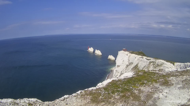 A panoramic view of the white chalk cliffs of a coastal landscape, with a lighthouse and a small building situated on top. The cliffs extend into the distance, with the ocean stretching out beneath a partly cloudy sky. Rock formations and greenery are visible along the edge of the cliffs.