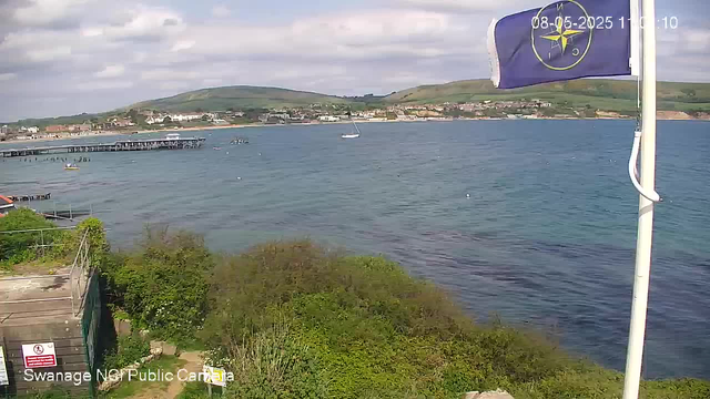 A view of a harbor with water and distant hills under a partly cloudy sky. A boat is sailing across the water, and there is a pier on the left. In the top right corner, a flag with a compass logo and a date and time stamp are visible. In the bottom left corner, there is signage that says "Swanage NCP Public Car" and something else partially obscured by foliage. Green bushes and a pathway are in the foreground.
