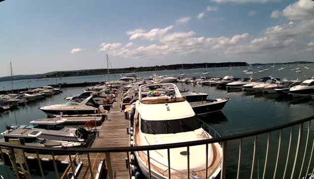 A view of a marina with several boats docked along a wooden pier, extending into the water. The sky is partly cloudy, and distant land is visible on the horizon. A black metal railing curves into the foreground, framing the scene.