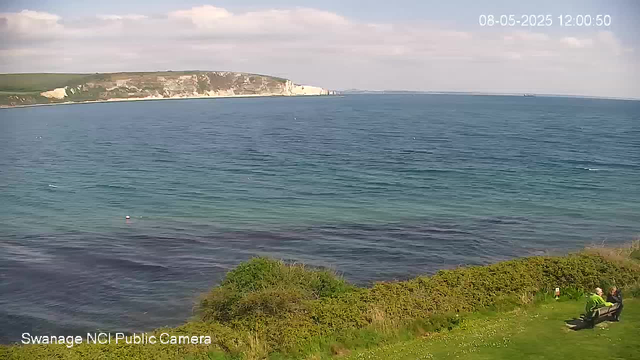A view of the ocean with gentle waves, a distant white chalk cliff on the horizon, and a partly cloudy sky. In the lower right corner, there are two people sitting on a bench, engaging in conversation. Green bushes and grass surround the seating area. The timestamp in the upper right corner shows the date and time as August 5, 2025, at 12:00:50. The bottom left corner indicates the camera is from Swanage NCI Public Camera.